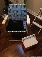 Front diagonal view of vintage metal lawn chair with blue and green plastic webbing, white armrests, black Gel pad on seat, and red/white Igloo cooler in front on wood floor.