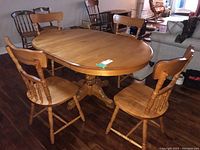 Full view of the oval wooden dining table with pedestal base, two extension leaves included, and four wooden chairs surrounding it for scale (chairs not included). Table shows minor wear but generally in good condition.
