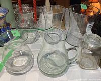 Group shot of 4 clear glass vases, 1 clear glass jar with lid, and crystal pitcher with green tint on table.