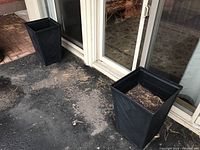 Two tall black resin planters placed outdoors near glass doors, containing soil and showing a geometric textured pattern.