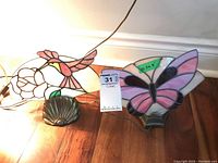 Two stained glass lamps on wooden floor near wall baseboard. One lamp depicts a hummingbird and flower design, the other a butterfly design. Both have metal shell-shaped bases and cords attached but bulbs not installed.
