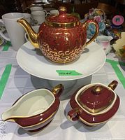 Teapot, creamer, sugar bowl on top of the white pedestal cake stand. The red and gold teapot is round with gold handle and spout, lid with knob. Creamer and sugar bowl are similarly decorated with gold trim.