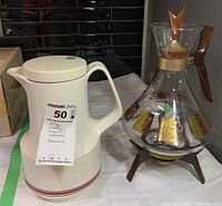 Full view of the white Thermos brand thermos with handle and the vintage Inland Glass coffee carafe with wooden handle and decorative gold pattern