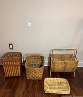 Four wicker items including a picnic basket with handle, large basket with handle, wicker magazine rack with wooden legs, and small flat tray arranged on wood floor against a wall