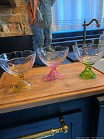 Close-up of three glass stemmed bowls with yellow, pink, and green stems and bases on a wooden surface, showing scalloped rims and textured bowls.
