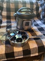 Instant Pot electric pressure cooker and stainless steel 6-egg poacher pan with glass lid placed on checkered fabric.