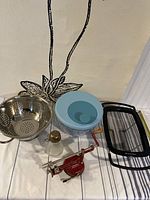 View of two colanders, an apple core peeler, an antique walnut jar, and a salad spinner on a table against a wall with decorative drawing.