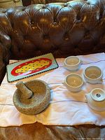 Set of kitchen serving pieces on a cloth background includes a colorful rectangular platter, four white ceramic soup bowls, heavy stone guacamole bowl with pestle.