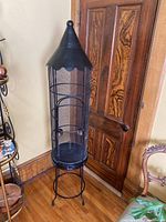 Full view of the tall, round metal bird cage with a pointed roof, standing on four legs.