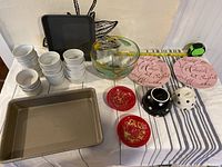 Photo showing a collection of kitchen items including a 9x9 Wilton baking pan, stacks of white ceramic custard dishes, a large glass bowl with painted fruit, two pink plates with script design, two red plates with gold details, black and white polka dot bowls, and gold speckled sushi dishes, all placed on a white tablecloth with blue stripes.