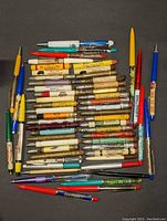 Top-down photo showing the full collection of approximately 45 vintage advertising bullet pencils and floaty pens on a black background, organized in neat rows. Varied colors and wear visible.