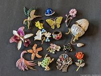 Top-down view showing an assortment of enamel brooches featuring various animals, flowers, and shapes including a hummingbird, butterfly, ladybug, frog, flowers, basket, and rose.