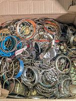 Box of assorted metal costume jewelry showing bangles, bracelets, necklaces, and earrings with various colors and designs piled together.