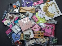 Photo showing a full assortment of costume jewelry in retail packaging including earrings, necklaces, bracelets, pins and pendants spread on a gray surface.