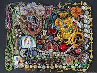 Full overhead view of the collection of colorful whimsical costume jewelry showing various necklaces, bracelets, earrings, pendants, and brooches in mixed materials and bright colors scattered on a black surface.