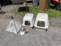 Wide shot showing all items in the lot: two litter bins with lids, small fabric cat tent, litter scoop and self-water feeder sitting outside on gravel ground.