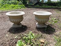 Two large round cement garden urns placed on dirt ground with grass and plants nearby, showing front and side details