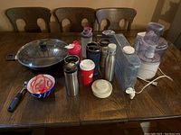 Photo of large wok with assorted water bottles, thermoses, cups, plates, strainers, and electric food processor on wooden table.