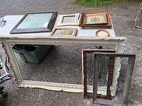 Large white distressed wooden mirror frame with a sizeable rectangular mirror leaning against a table under natural light outdoors, showing wear consistent with vintage or shabby chic style.
