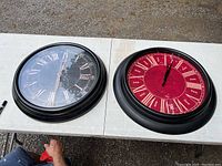 Two large round wall clocks side by side on a white table, black clock on left (23 inches), red clock on right (25 inches). Both have Roman numerals and glass covers.