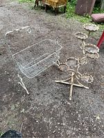 Photo showing decorative white metal cradle with attached curved handle and wire basket beside a multi-tier metal plant stand with round scalloped shelves on a dirt ground outdoors.