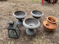 Photo showing three plastic grey garden urns with fluted design, terra-cotta herb pot, black metal lantern, and rust-colored ash bin all arranged on dirt ground.