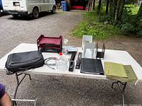 Wide shot of entire lot on a white folding table outdoors including computer bag, burgundy stackable desk organizer, candy dish, hole punch, stapler, paperwork holders, DayTimer planner, folders, and tape holder.