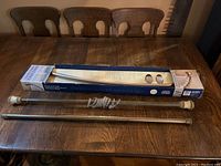 Wide shot showing two shower curtain rods, one boxed and one unboxed, on wood table surface, with mounting hardware laid out.