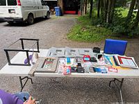 Photo of entire lot on white folding table outdoors showing various office supplies including two file racks, paper items, stamp pads, and holders.