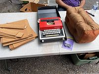 Photo showing red manual typewriter in grey carrying case with new ribbon cartridge, stack of cork sheets to left, and rust orange decorative pillow to right on a white folding table.