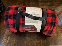 Rolled weighted throw blanket with red and black checked pattern, secured by strap, showing the product label.