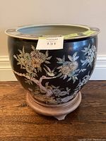Front view of the black ceramic planter showing floral and bird motifs and wooden stand beneath.