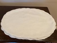 Stack of 12 white oval scalloped placemats on dark wood surface