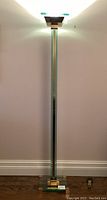 Full view of the tall clear glass and brass floor lamp illuminated against a wall, showing the entire lamp height and base.