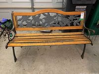 Front view of outdoor bench showing wooden seat slats, painted wooden top edge, and metal floral backrest with roses and leaves patterns.