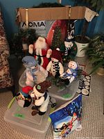 View of the holiday figurines including Santa, snowmen, penguin placed on clear plastic storage bins with a glimpse of the Noma Christmas tree box in the background