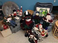 Multiple Christmas figurines and stuffed characters arranged on floor and chairs including snowmen, Santas and reindeer.
