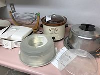 Overview of all items in lot showing crock pot, electric grill, fruit basket, cake preserver, cake carrier, and splash guard on countertop