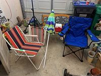 Two metal and plastic striped folding chairs, blue folding captain chair with cup holder, beach umbrella with blue and green stripes, small blue Coleman cooler. Items shown in a storage or garage setting.