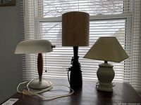 Three table lamps on a wooden surface in front of a window with blinds. From left: a desk lamp with frosted glass shade and wooden base, a wooden lamp with cylindrical base and textured beige shade, and a white ceramic lamp with a conical beige shade.