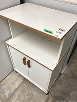 Front view of white laminate microwave stand with wooden edge trim, open middle shelf, and lower cabinet with two wooden handle doors.