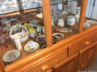 View of a shelf with multiple small plates and vases including Easter and ceramic items visible through glass cabinet doors