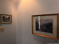 Two framed landscape prints hanging on white walls including one in an ornate gold frame with a rustic scene and another smaller with a cabin and lake scene.