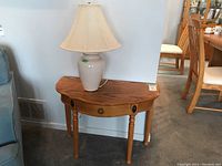 Demi lune wood veneer side table with single drawer and ceramic lamp on top, showing overall wood finish and lamp details.