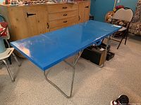 Blue painted metal folding table fully extended on a carpeted floor in a room with wooden drawers.