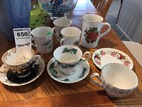 Photo of multiple teacups, saucers, and mugs arranged on wooden table. Shows diversity of patterns and styles including black and gold ornate cup, floral prints, and bird design.