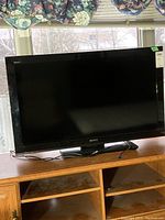 Front view of Sony Bravia TV showing full screen and stand on wood cabinet beneath window with floral curtain blinds.