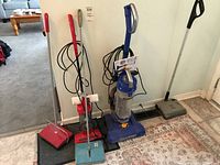 Photo of the Eureka and Dirt Devil vacuums alongside three manual carpet sweepers arranged against a wall, showing all items in the lot.