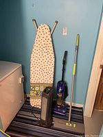 Full view of vacuum cleaner, heater, ironing board, and mop against blue wall.