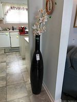 Full view of tall black ceramic floor vase with faux orchid stem placed in the interior corner of a room near a kitchen.